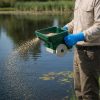 Blue nitrile glove uses green Ironvale hand spreader to apply granular aquatic herbicide/algaecide along weeded shoreline; tan cuff.