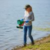 Female professional applicator wearing long sleeves and gloves applying gray granular treatment into lake along shoreline with handheld spreader.