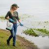 Female professional applicator wearing long sleeves and gloves spraying aquatic product into pond along shoreline with handheld pump sprayer.