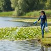 Female Pro applicator wearing long sleeves and gloves spraying aquatic product into pond along shoreline with handheld pump sprayer.