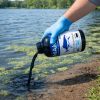 Gloved hand pouring MAKO 5x Concentrate Dye from the bottle along a pond shoreline