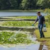 Male professional applicator wearing long sleeves and gloves spraying aquatic liquid into pond along shoreline with handheld Ironvale pump sprayer.