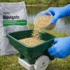 Tan Navigate Plus granules poured from a clear cup into a green Ironvale hand spreader beside a pond