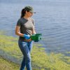 Female professional applicator wearing short sleeves and gloves applying blue granular treatment into lake along shoreline with handheld spreader.